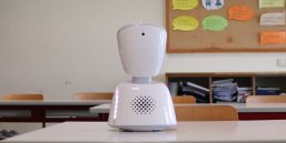 Avatar in the classroom A small white robot stands on a brown table in a classroom.