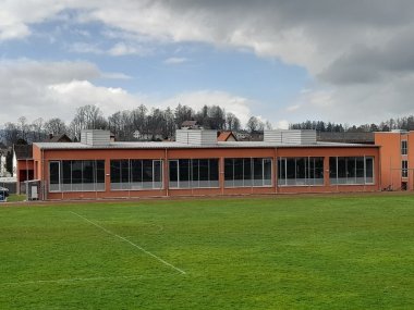 Photo of the exterior facade of the Bad Tölz grammar school gymnasium. In the foreground is the lawn of the sports field. The hall is red, single-storey and has many windows. The sky is gray and cloudy.