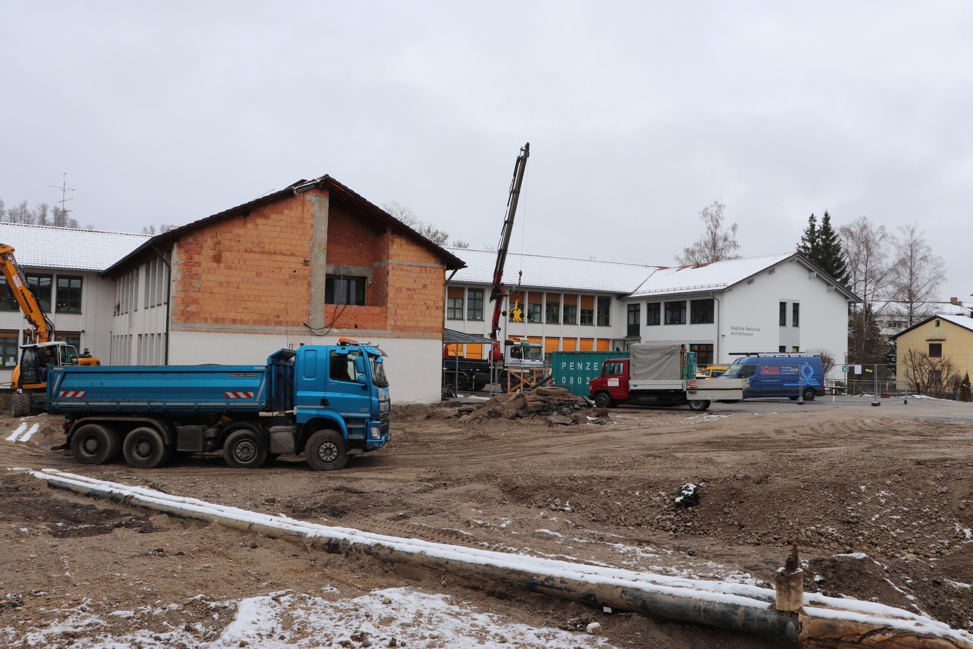 The picture shows the Wolfratshausen secondary school in the background. A blue truck passes in front of the building. In the foreground, two pipes can be seen running along the ground from the left center of the picture to the bottom right. At the bottom right, the ground sinks down to a pit.