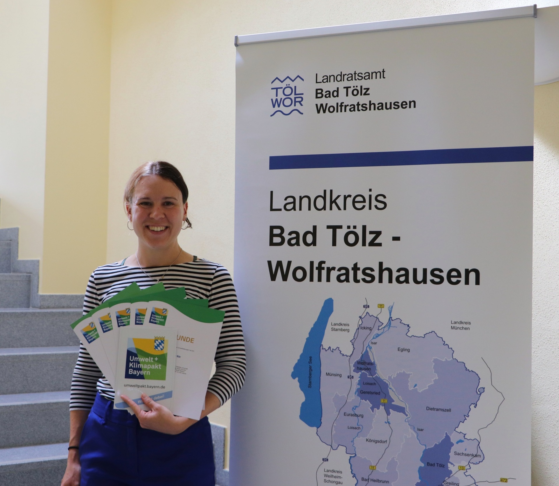 Climate Protection Manager Veronika Böhm holds the certificates for the new members of the Bavarian Environmental and Climate Protection Pact. She is standing next to a roll-up. The map of the district is depicted on the roll-up.