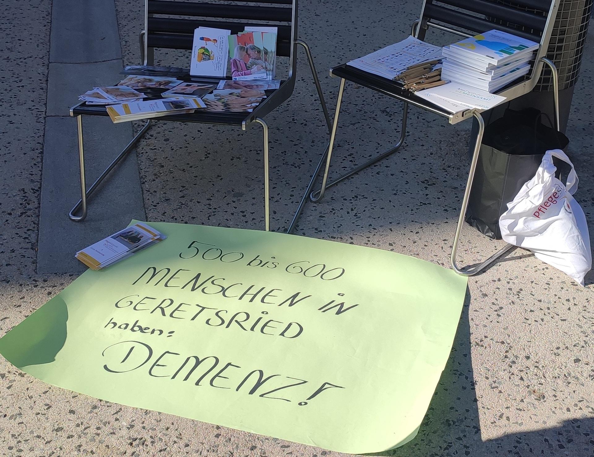 A poster lies on the floor; it reads, in black letters on a green background: “500 to 600 people in Geretsried have dementia!” Behind the poster are two chairs. A bag bearing the logo of the care center is leaning against the chair on the right.