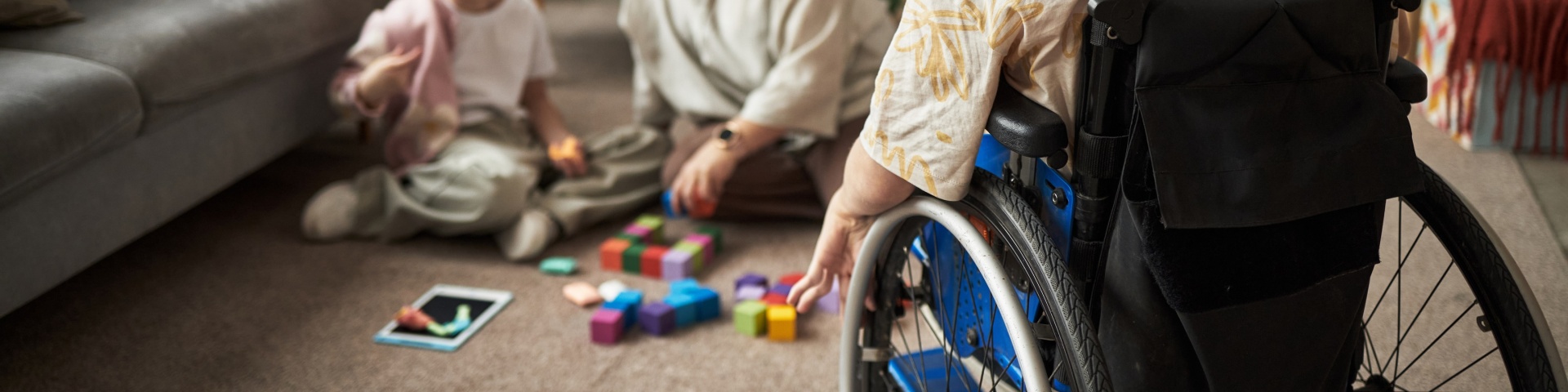 A youngster in a wheelchair sits on the carpet while another child plays on the floor with an adult.