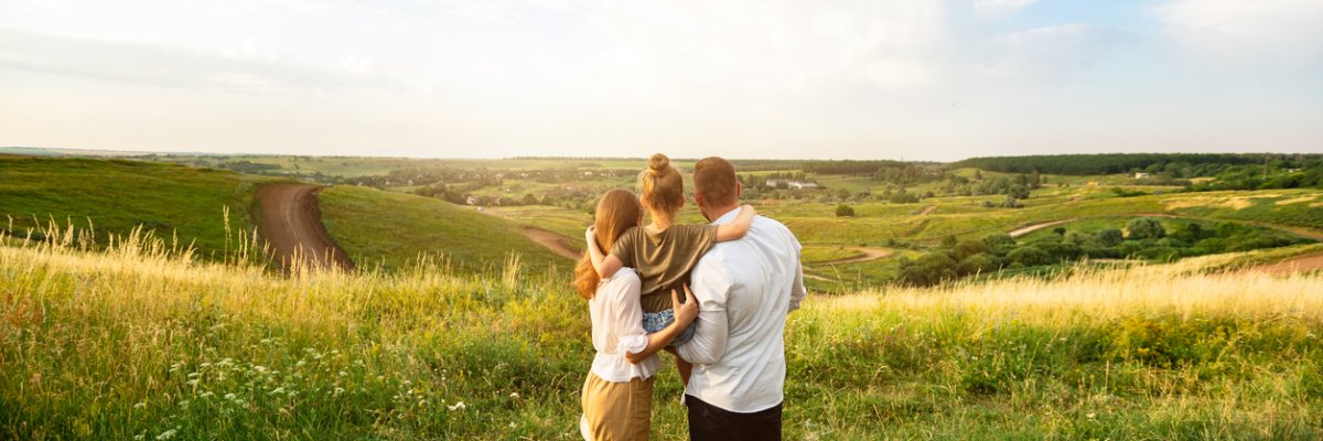The picture shows a family with a father, mother and child. They are standing on a country lane, gazing together into the distance over a vast, idyllic landscape.