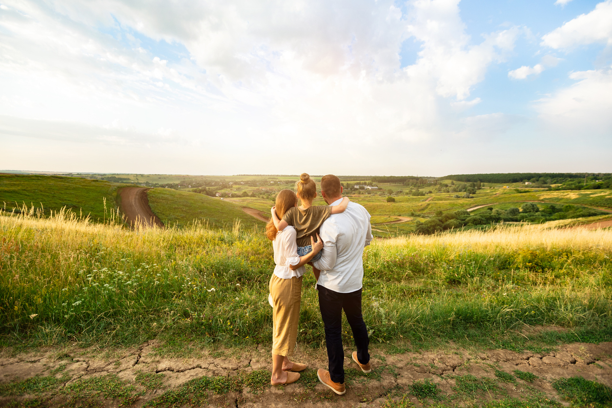 The picture shows a family with a father, mother and child. They are standing on a country lane, gazing together into the distance over a vast, idyllic landscape.