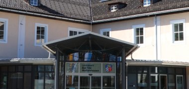 The picture shows the entrance area of the district administration office. View of the inner courtyard from the center of the traffic circle. You can see a tin roof supported by red columns. The glass façade and sliding entrance door are in the background. The building is apricot-colored.