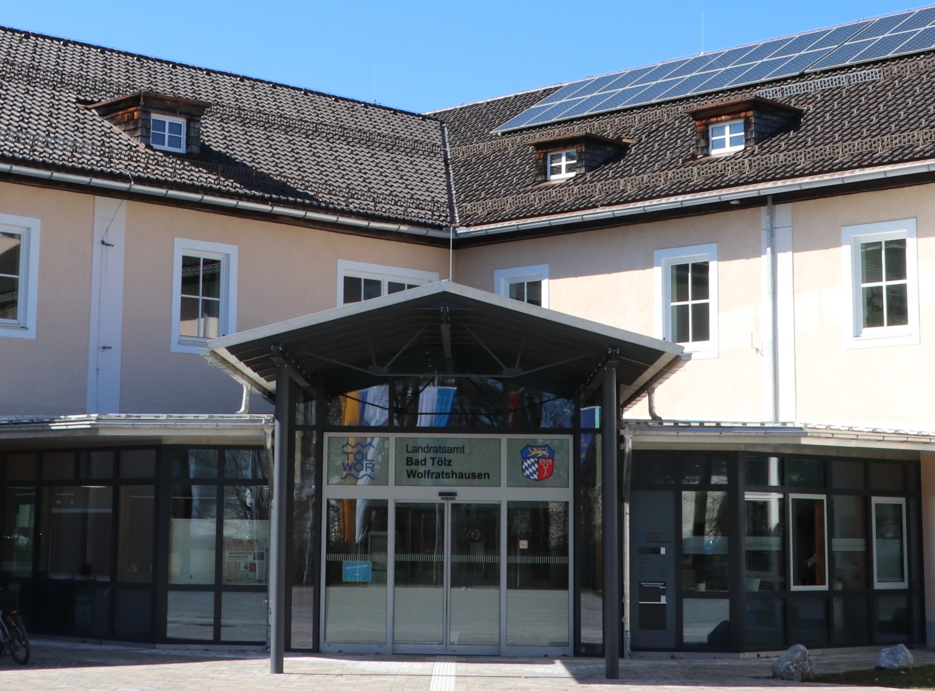The picture shows the entrance area of the district administration office. View of the inner courtyard from the center of the traffic circle. You can see a tin roof supported by red columns. The glass façade and sliding entrance door are in the background. The building is apricot-colored.