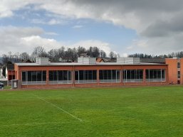 Foto der Aussenfassade der Turnhalle des Gymnasium Bad Tölz. Foto der Aussenfassade der Turnhalle des Gymnasium Bad Tölz. Im Vordergrund der Rasen des Sportplatzes. Die Halle ist rot, einstöckig und hat viele Fenster. Der Himmel ist grau und bewölkt.