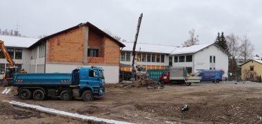 Realschule Wolfratshausen: Die Vorbereitungen für die Baugrube laufen. Das Bild zeigt im Hintergrund die Realschule Wolfratshausen. Vor dem Gebäude fährt ein blauer Laster vorbei. Im Vordergrund sieht man zwei Rohre, die auf dem Erdreich von der linken Bildmitte nach rechts unten verlaufen. Rechts unten senkt sich das Erdreich zu einer Grube hinab.