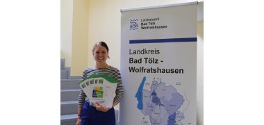 Klimaschutzmanagerin Veronika Böhm hat die Urkunden für die neuen Mitglieder im Umwelt- und Klimaschutzpakt Bayern in der Hand. Sie steht neben einem Rollup. Auf dem Rollup ist die Landkreiskarte abgegebildet.