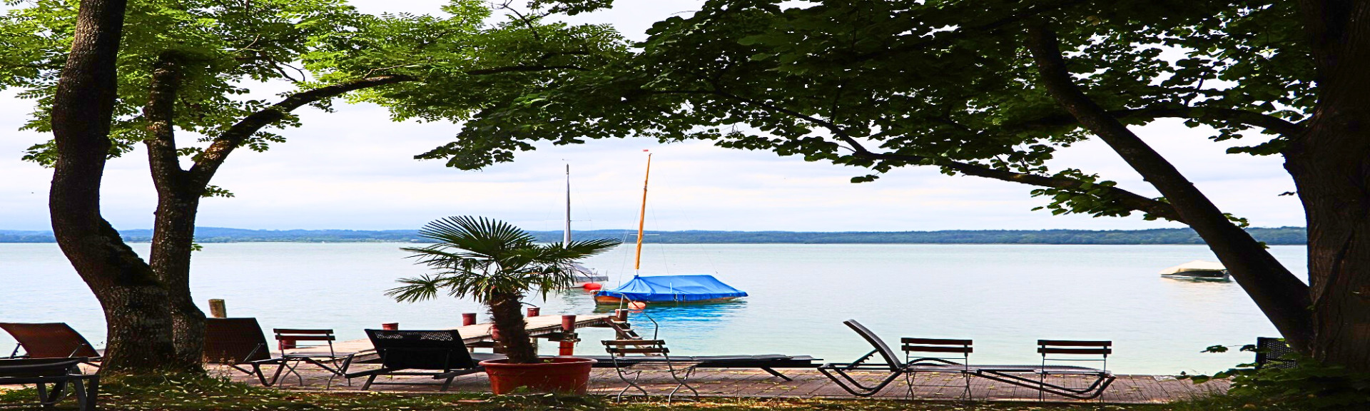 Blick vom Gras-Badestrand über Holzdeck und  Badesteg auf das Wasser des Starnberger Sees. Links und Rechts Laubbäume. Auf dem Wasser mit Planen abgedeckte Segelboote. Am Strand Strandmöbel aus Holz und Metall. Man sieht das Ufer der anderen Seite im Dunst.