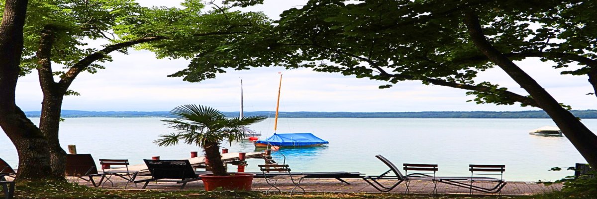Blick vom Gras-Badestrand über Holzdeck und  Badesteg auf das Wasser des Starnberger Sees. Links und Rechts Laubbäume. Auf dem Wasser mit Planen abgedeckte Segelboote. Am Strand Strandmöbel aus Holz und Metall. Man sieht das Ufer der anderen Seite im Dunst.