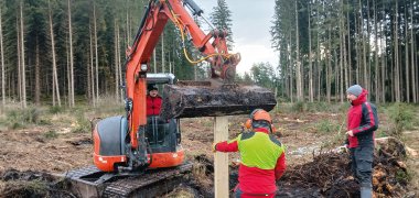 Aufnahme mit Moorboden vor Wald, ein BAgger und zwei Männer in Arbeitsuniform