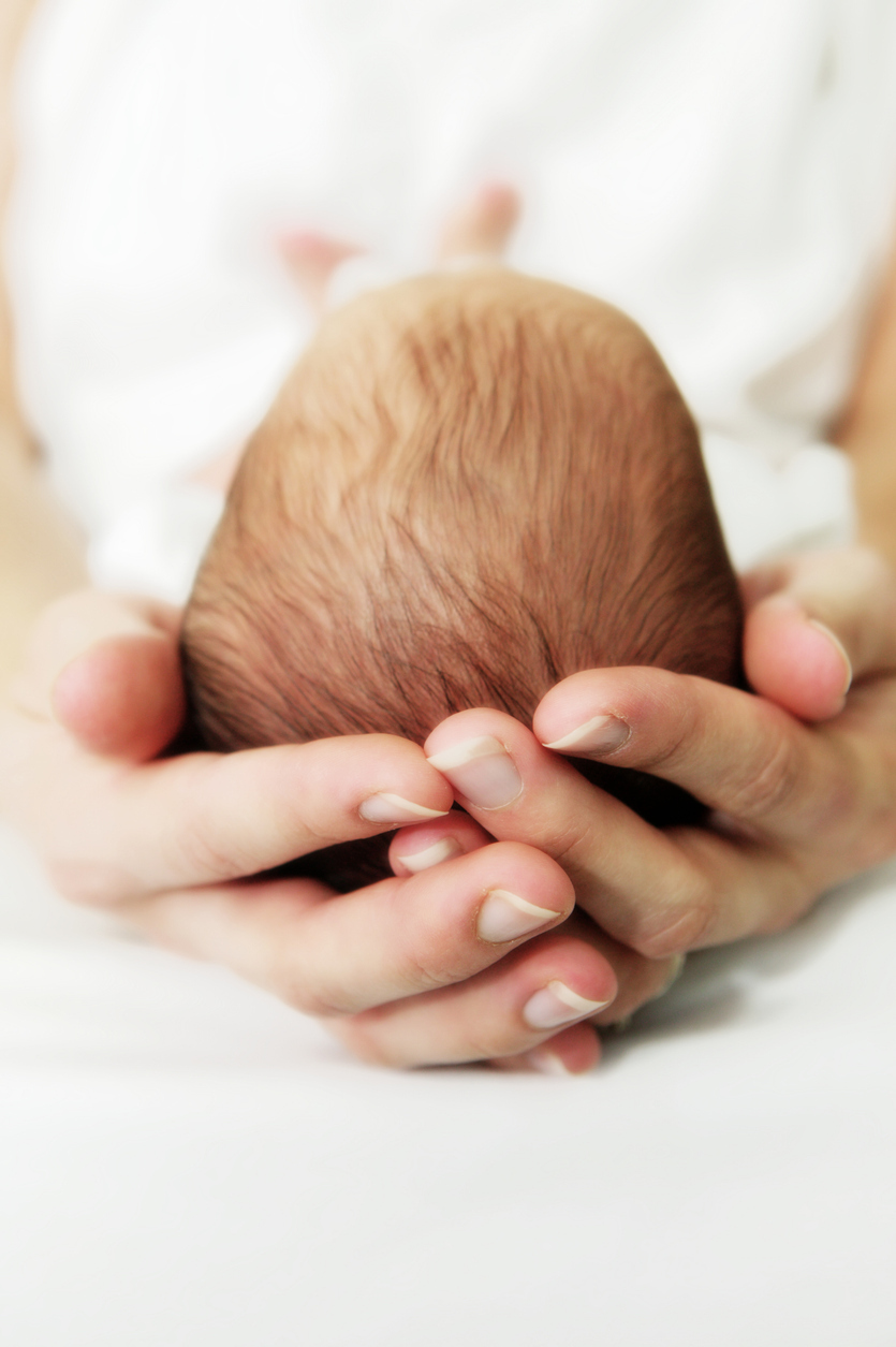Ein Erwachsener hält den Kopf eines Babys vorsichtig in beiden Händen. Man sieht nur den Hinterkopf des Babys mit feinen Haaren.