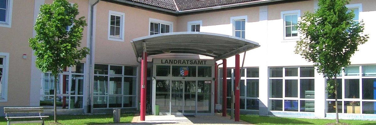 Eingang des Landratsamts Auf dem Bild dargestellt ist der Eingangsbereich des Landratsamts. Sichtwinkel Innenhof aus der Mitte des Rondells. Sie sehen ein Blechdach getragen von roten Säulen. Im Hintergrund die Glasfassade und Eingangsschiebetür. Das Gebäude ist Aprikotfarben.