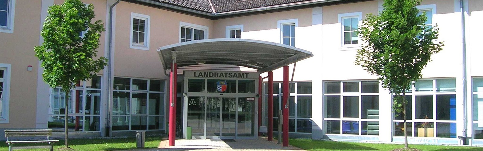 Auf dem Bild dargestellt ist der Eingangsbereich des Landratsamts. Sichtwinkel Innenhof aus der Mitte des Rondells. Sie sehen ein Blechdach getragen von roten Säulen. Im Hintergrund die Glasfassade und Eingangsschiebetür. Das Gebäude ist Aprikotfarben.