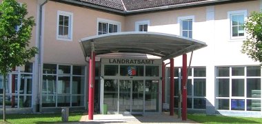 Eingang des Landratsamts Auf dem Bild dargestellt ist der Eingangsbereich des Landratsamts. Sichtwinkel Innenhof aus der Mitte des Rondells. Sie sehen ein Blechdach getragen von roten Säulen. Im Hintergrund die Glasfassade und Eingangsschiebetür. Das Gebäude ist Aprikotfarben.