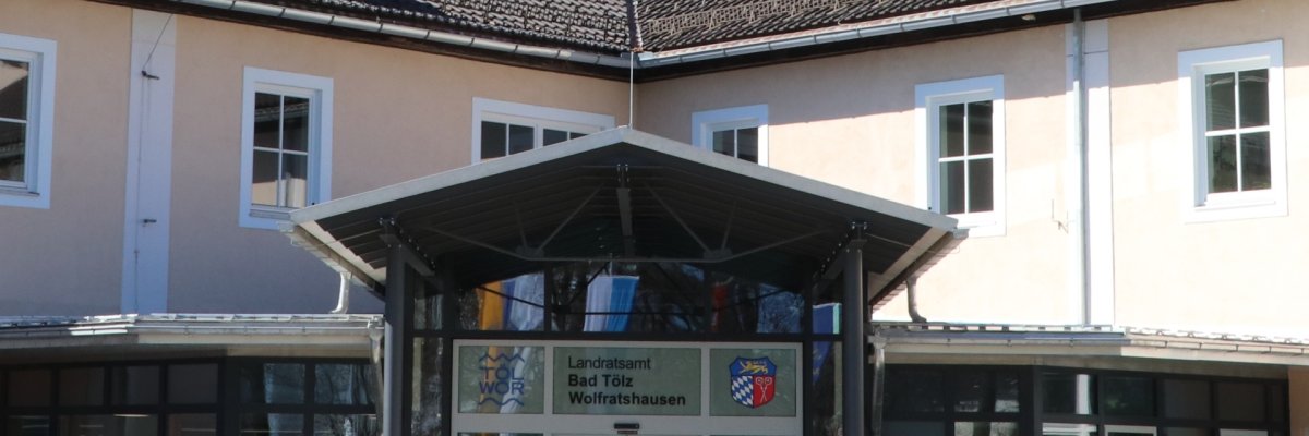 Auf dem Bild dargestellt ist der Eingangsbereich des Landratsamts. Sichtwinkel Innenhof aus der Mitte des Rondells. Sie sehen ein Blechdach getragen von roten Säulen. Im Hintergrund die Glasfassade und Eingangsschiebetür. Das Gebäude ist Aprikotfarben.